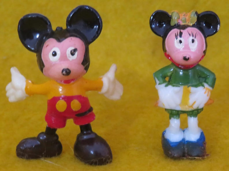 May include: Two small, vintage Mickey Mouse and Minnie Mouse figurines. Mickey is in a yellow and red outfit with black ears and shoes. Minnie is in a green top, white skirt, and blue shoes, with a bow on her head. Both have pink faces.