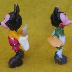 May include: Two small, vintage figurines of cartoon characters. One figure is dressed in red pants, a yellow shirt, and black shoes. The other figure is wearing a green top, white pants, and blue shoes. Both have black ears and pink faces.