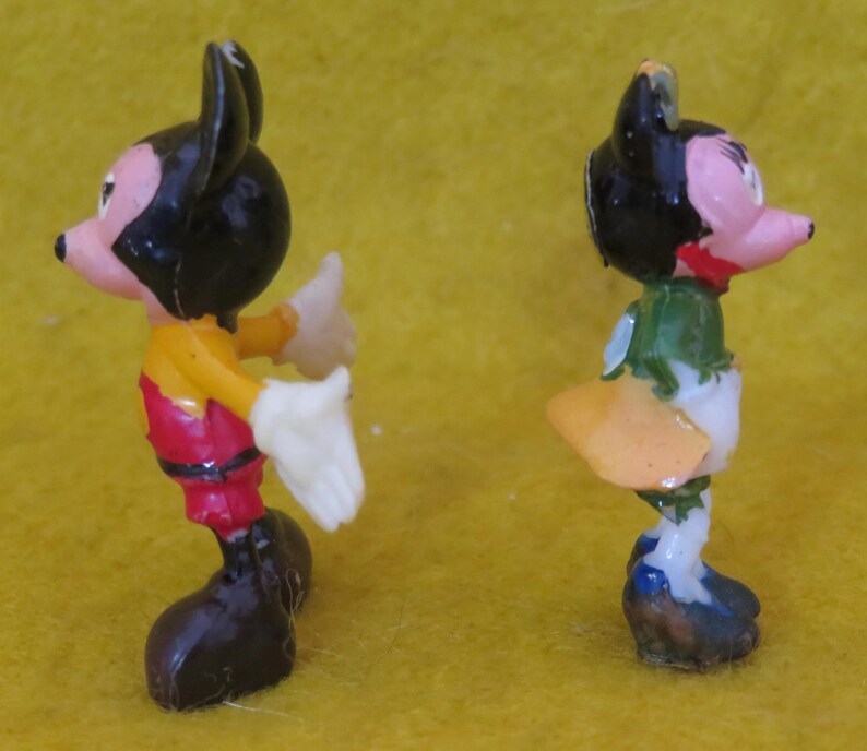 May include: Two small, vintage figurines of cartoon characters. One figure is dressed in red pants, a yellow shirt, and black shoes. The other figure is wearing a green top, white pants, and blue shoes. Both have black ears and pink faces.