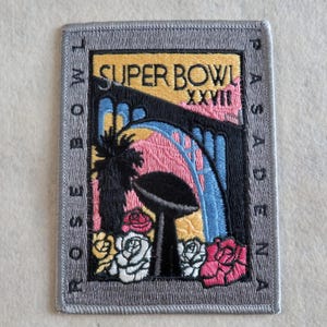May include: A rectangular embroidered patch with a gray border featuring the text SUPER BOWL XXVII, ROSE BOWL, and PASADENA. The design includes a black trophy, a palm tree, blue arches, and pink and white roses against a yellow and black background.