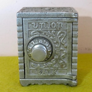 May include: A silver-colored, vintage Union Bank safe box. The safe box has a combination dial with the numbers 7, 8, and 9 visible. The word "UNION" is at the top, and "BANK" is at the bottom. The safe box has an ornate design.