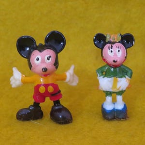 May include: Two small figurines of Mickey and Minnie Mouse. Mickey is wearing a yellow shirt, red shorts, and black shoes, with his arms raised in a thumbs-up gesture. Minnie is in a green top, white skirt, and blue shoes.