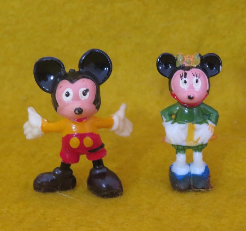 May include: Two small figurines of Mickey and Minnie Mouse. Mickey is wearing a yellow shirt, red shorts, and black shoes, with his arms raised in a thumbs-up gesture. Minnie is in a green top, white skirt, and blue shoes.