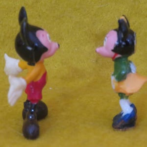 May include: Two small figurines of cartoon characters. One figure is wearing a yellow shirt, red overalls, white gloves, and black shoes. The other figure is wearing a green jacket, orange skirt, and blue shoes. Both figures have black hair and pink faces.