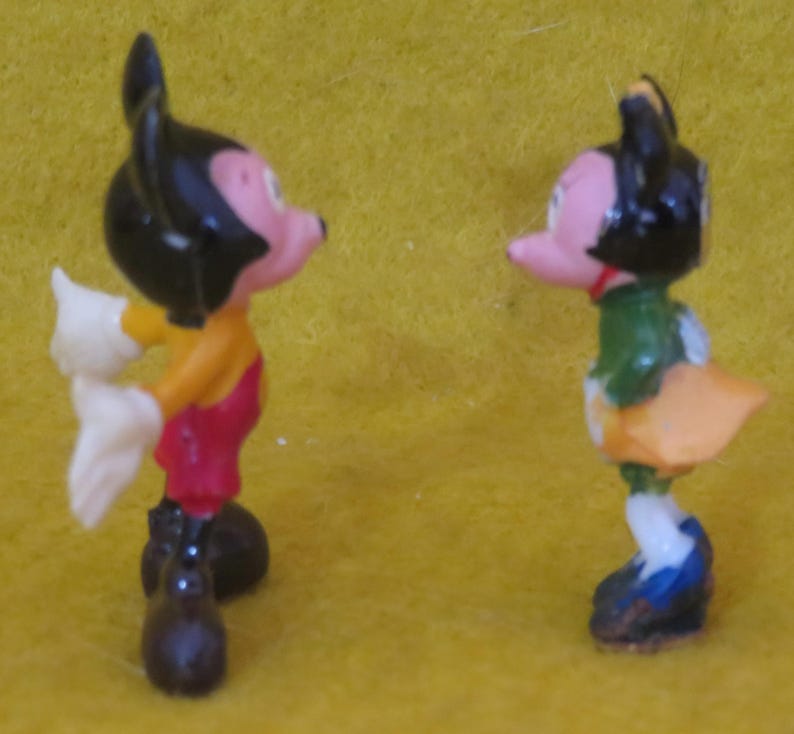 May include: Two small figurines of cartoon characters. One figure is wearing a yellow shirt, red overalls, white gloves, and black shoes. The other figure is wearing a green jacket, orange skirt, and blue shoes. Both figures have black hair and pink faces.