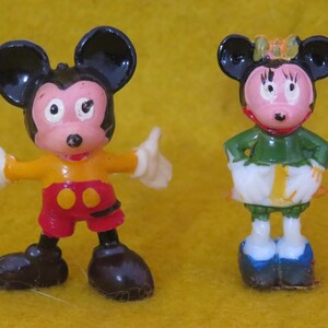 May include: Two vintage Mickey Mouse and Minnie Mouse figurines. Mickey is wearing a yellow shirt, red shorts, and black shoes. Minnie is wearing a green top, white skirt, and blue shoes. Both figures have black ears and are standing on a yellow surface.