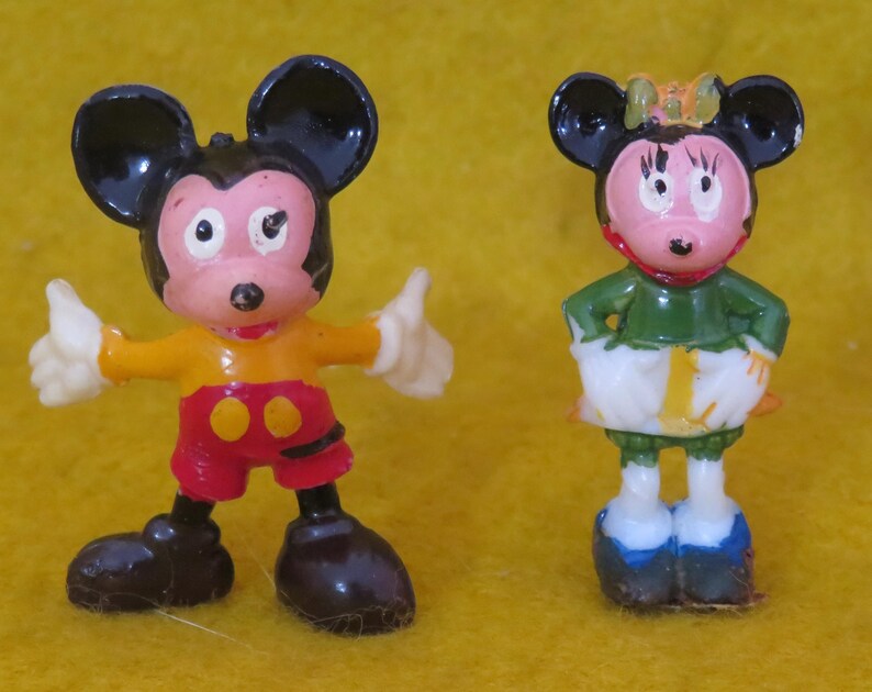May include: Two vintage Mickey Mouse and Minnie Mouse figurines. Mickey is wearing a yellow shirt, red shorts, and black shoes. Minnie is wearing a green top, white skirt, and blue shoes. Both figures have black ears and are standing on a yellow surface.