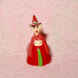 May include: A small, red and green painted figurine of a fairy wearing a pointed hat with a blue and white design. The fairy has a painted face with a smile and large eyes.