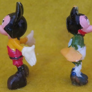 May include: Two small, vintage figurines of cartoon characters. One figure is Mickey Mouse, wearing red shorts, a yellow shirt, and black shoes. The other figure is Donald Duck, wearing a blue and white outfit with a yellow tail. Both figures are on a yellow background.