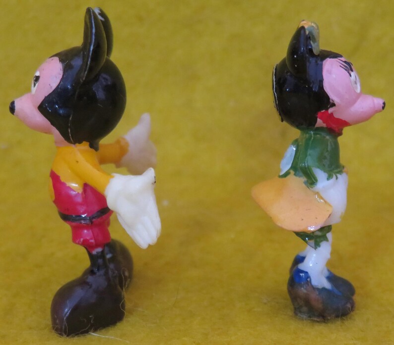 May include: Two small, vintage figurines of cartoon characters. One figure is Mickey Mouse, wearing red shorts, a yellow shirt, and black shoes. The other figure is Donald Duck, wearing a blue and white outfit with a yellow tail. Both figures are on a yellow background.