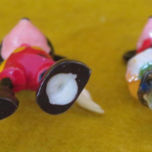 May include: Two small, colorful Mickey Mouse figurines. One is lying on its back, showing red shorts, black shoes, and a yellow tail. The other figurine is missing parts, with a pink head and a colorful outfit.