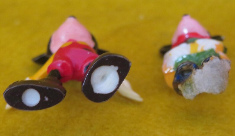 May include: Two small, colorful Mickey Mouse figurines. One is lying on its back, showing red shorts, black shoes, and a yellow tail. The other figurine is missing parts, with a pink head and a colorful outfit.