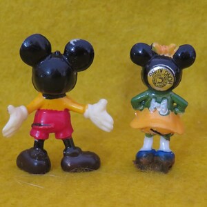 May include: Two small Mickey Mouse figurines. One figure is Mickey Mouse, wearing red shorts, yellow shirt, black shoes, and white gloves. The other figure is Minnie Mouse, wearing a green top, orange skirt, and blue shoes. Both figures have black ears.