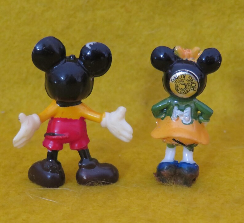 May include: Two small Mickey Mouse figurines. One figure is Mickey Mouse, wearing red shorts, yellow shirt, black shoes, and white gloves. The other figure is Minnie Mouse, wearing a green top, orange skirt, and blue shoes. Both figures have black ears.