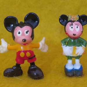 May include: Two small figurines of cartoon mice. One mouse is wearing a yellow shirt, red shorts, and black shoes, with white gloves and black ears. The other mouse is wearing a green top, white skirt, and blue shoes, with a bow and black ears.