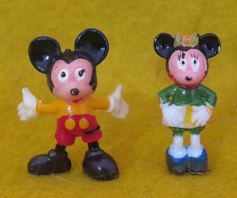 May include: Two small figurines of cartoon mice. One mouse is wearing a yellow shirt, red shorts, and black shoes, with white gloves and black ears. The other mouse is wearing a green top, white skirt, and blue shoes, with a bow and black ears.