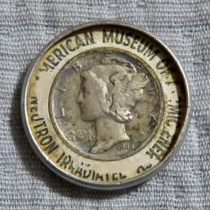 May include: A silver-colored coin with the text "AMERICAN MUSEUM OF MICHEL" and "NEUTRON IRRADIATED" around the edge. The coin features a profile of a woman with a winged cap and the year 1944.