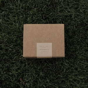 May include: A rectangular brown cardboard box featuring a centred paper band with the printed text "AMONG THE FLOWERS" and the handwritten-style word "summer" in black ink, providing a simple and clean presentation for the enclosed item.