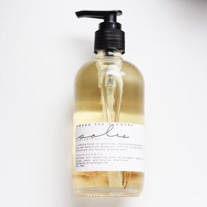 May include: A clear glass bottle with a black pump dispenser filled with a light yellow liquid. The bottle has a white label with the text "AMONG THE FLOWERS solis RESTORATION OIL A  livening blend of uplifting, mood boosting essential oils and reparative botanical oils for relief from afflictions and healthy glowing skin. INGREDIENTS sunflower oil, essential oils of bergamot, lime, orange, lemon, and vanilla, starflower.  DISTRIBUTED IN CALIFORNIA USA."