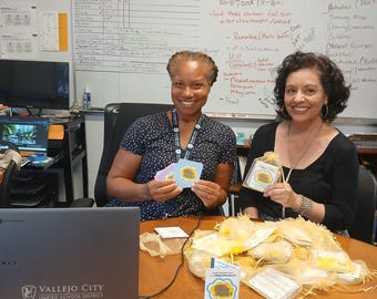 May include: Two women seated at a table with a laptop and various items. One woman holds cards with a blue and yellow design, while the other holds small bags. A sign on the table reads "Happy Prescription". A whiteboard is in the background.