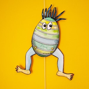 May include: A whimsical, hand-painted paper puppet of an egg-shaped creature with blue and green stripes, blue legs, and spiky hair. The puppet has large eyes and is suspended by a yellow string against a bright yellow background. The website wi-la-ne.de is printed on the puppet.