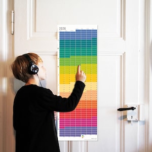 May include: A colorful 2026 wall calendar with a rainbow gradient design. The calendar is rectangular and mounted on a white door. The person in the image is wearing headphones and pointing at the calendar.