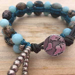 May include: Handmade bracelet featuring a mix of blue, brown, and silver beads. The bracelet is crafted with a teal cord and leather straps. A silver button with a world map design serves as a clasp. The bracelet is approximately 7 inches in circumference.