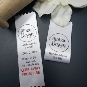 May include: Two white satin ribbon labels with black text and a black circle logo that says "Ribbon Design". The label on the left has additional text that says "Handmade in the UK", "100% Cotton", "Wash at 30c", "Low iron", "No tumble dry", and "KEEP AWAY FROM FIRE".