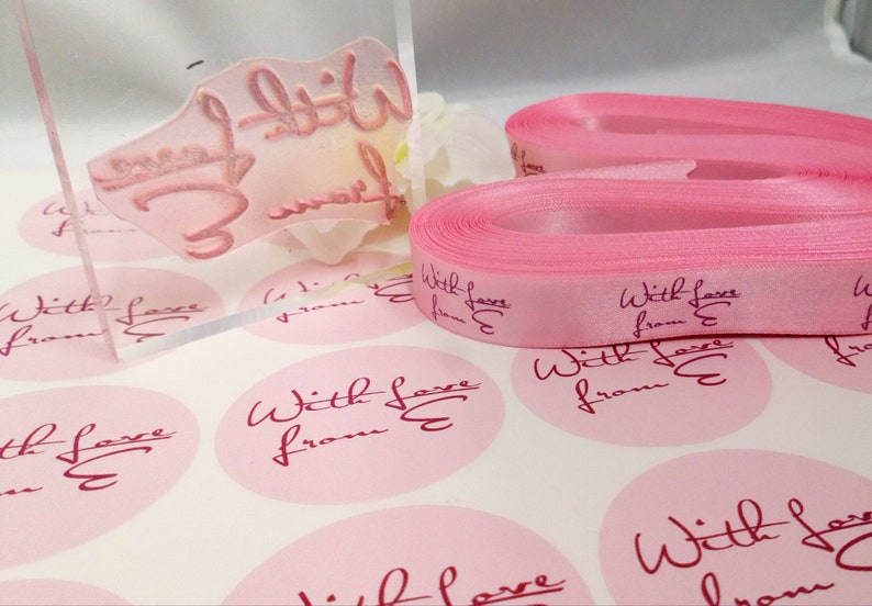 Puede incluir: Cinta de sat&eacute;n rosa con el texto "With Love from E" impreso en ella. Tambi&eacute;n hay varias pegatinas redondas rosas con el mismo texto.