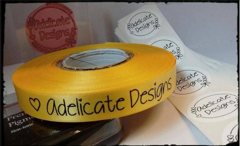 Puede incluir: Un rollo de cinta de sat&eacute;n amarilla con el texto "Adelicate Designs" impreso en negro. La cinta est&aacute; envuelta alrededor de un carrete y hay varias pegatinas blancas redondas con el mismo texto impreso en negro.