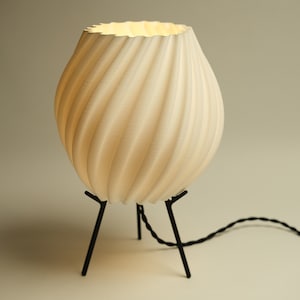 Op de afbeelding: Een witte, 3D-geprinte tafellamp met een draaiende, textuurrijke kap en een zwarte metalen driepoot.