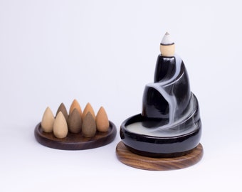 May include: A black ceramic incense burner with a cascading design, emitting white smoke. The burner sits on a wooden base, with a wooden tray holding several incense cones in shades of brown. A decorative and aromatic item.