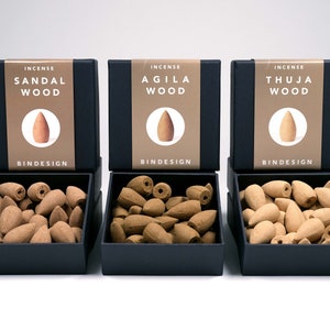 Op de afbeelding: Drie zwarte dozen met wierookkegels met de labels "Sandal Wood", "Agila Wood" en "Thuja Wood". Elke doos heeft een bruin label met een kegelillustratie. De dozen zijn open en tonen de wierookkegels.