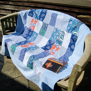 Peut inclure: Une courtepointe bleue et blanche à motifs floraux est drapée sur un banc en bois. Un petit livre au motif rouge et blanc repose sur la courtepointe.