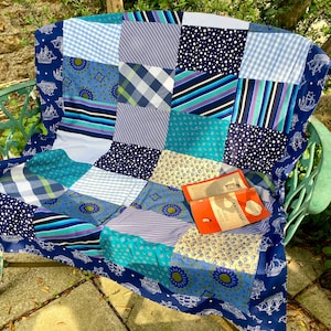 Op de afbeelding: Een patchwork quilt in blauwe en turquoise tinten, met diverse patronen, over een groene metalen bank. Een boek met een oranje kaft ligt erop. De rand van de quilt heeft een nautisch ontwerp.