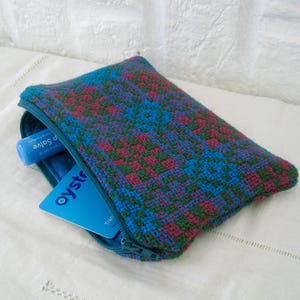 May include: A small, rectangular, zippered pouch with a woven pattern in teal, magenta, and green. The pouch is partially open, revealing a blue lip balm and a blue card with the word "oyster" printed on it.