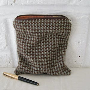 Puede incluir: Un estuche marrón y azul con estampado de pata de gallo y cremallera marrón. Un bolígrafo dorado y negro está junto al estuche. El estuche está hecho de tela texturizada.