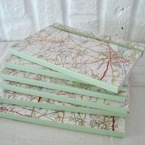 May include: A stack of six notebooks with map-printed covers. The edges of the notebooks are painted a pale green, and a green elastic band secures the top notebook. The maps show roads, towns, and other geographical details.