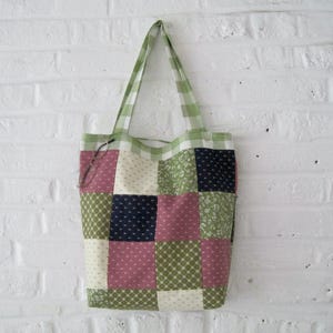 Pode incluir: Uma sacola tote de patchwork com uma alça e acabamento superior em xadrez verde e branco. O corpo da sacola é um patchwork de quadrados em tecidos rosa, verde, creme e azul marinho. A sacola está pendurada em uma parede de tijolos brancos.
