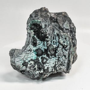May include: A dark grey mineral specimen with teal and black patches. The surface has a textured, bubbly appearance, with small, spherical formations. The overall shape is irregular and rough.