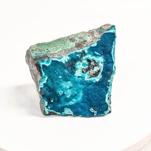 May include: A rough-cut mineral specimen with a vibrant display of colors. The stone features a deep teal blue hue with areas of turquoise and a band of green along the top edge. The edges are a dark gray, creating a striking contrast.
