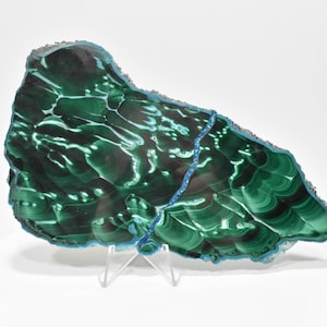 May include: A large, green malachite slab with a blue-green border. The slab is on a white stand and has a swirling, banded pattern.