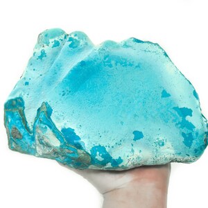 May include: A large, irregularly shaped turquoise blue stone with white and green markings. The stone is being held in a hand.
