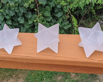 Puede incluir: Tres objetos transparentes en forma de estrella están dispuestos sobre una superficie de madera marrón. Los objetos tienen un diseño geométrico y parecen estar hechos de un material translúcido. El follaje verde es visible en el fondo.