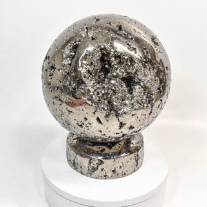 May include: A metallic silver sphere with a rough, textured surface and a large, open cavity filled with sparkling crystals. The sphere sits on a matching cylindrical base, creating a unique decorative object.