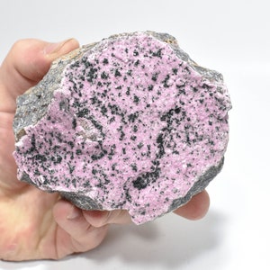 May include: A pink and black mineral specimen with a rough, textured surface. The mineral is a light pink colour with black specks throughout.