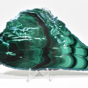 May include: A large, green malachite slab with a swirling, banded pattern. The slab is polished and has a smooth, glossy finish. It is displayed on a clear acrylic stand.