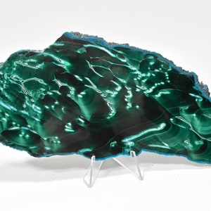 May include: A large, green malachite slab with a swirling, banded pattern. The slab is polished and has a smooth, glossy finish. It is displayed on a clear acrylic stand.