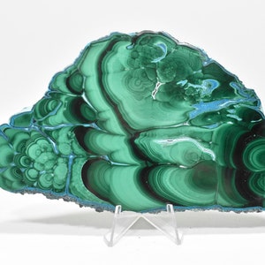 May include: A large, green malachite slab with swirling patterns and a dark green border. The slab is on a clear acrylic stand.