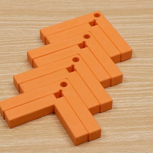 May include: Orange, L-shaped trivets arranged in a zig-zag pattern. Each trivet has a small hole in the corner. The trivets are made of a solid material and are resting on a light brown wooden surface.